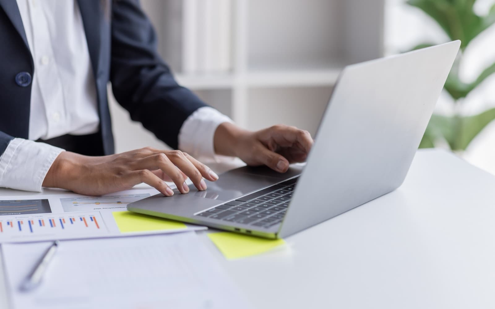 A person in a navy jacket typing on a laptop at a white desk, with printed charts beside it