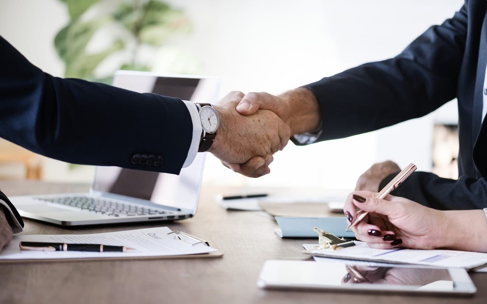 Two people in suits shaking hands across a desk, with a laptop and signed papers between them