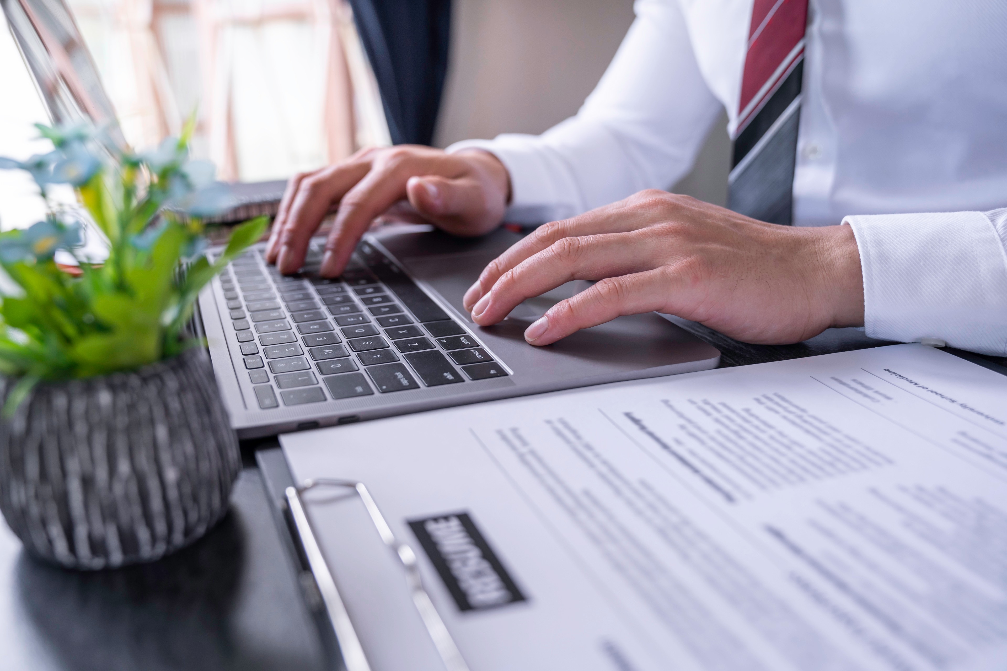 A person in a shirt and tie typing on a laptop at a desk, with a printed CV beside the laptop