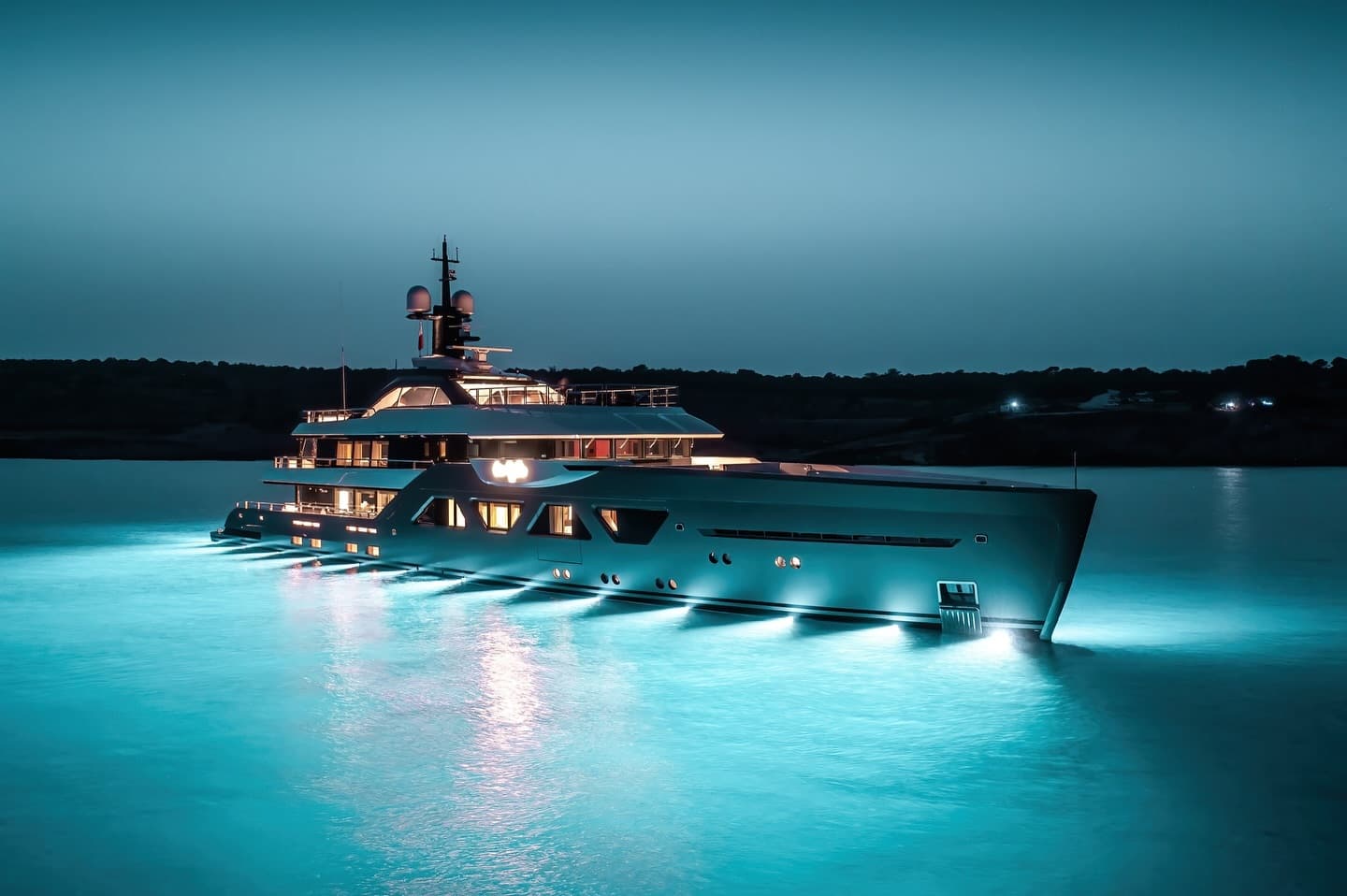 A superyacht at anchor at dusk with its underwater lights on