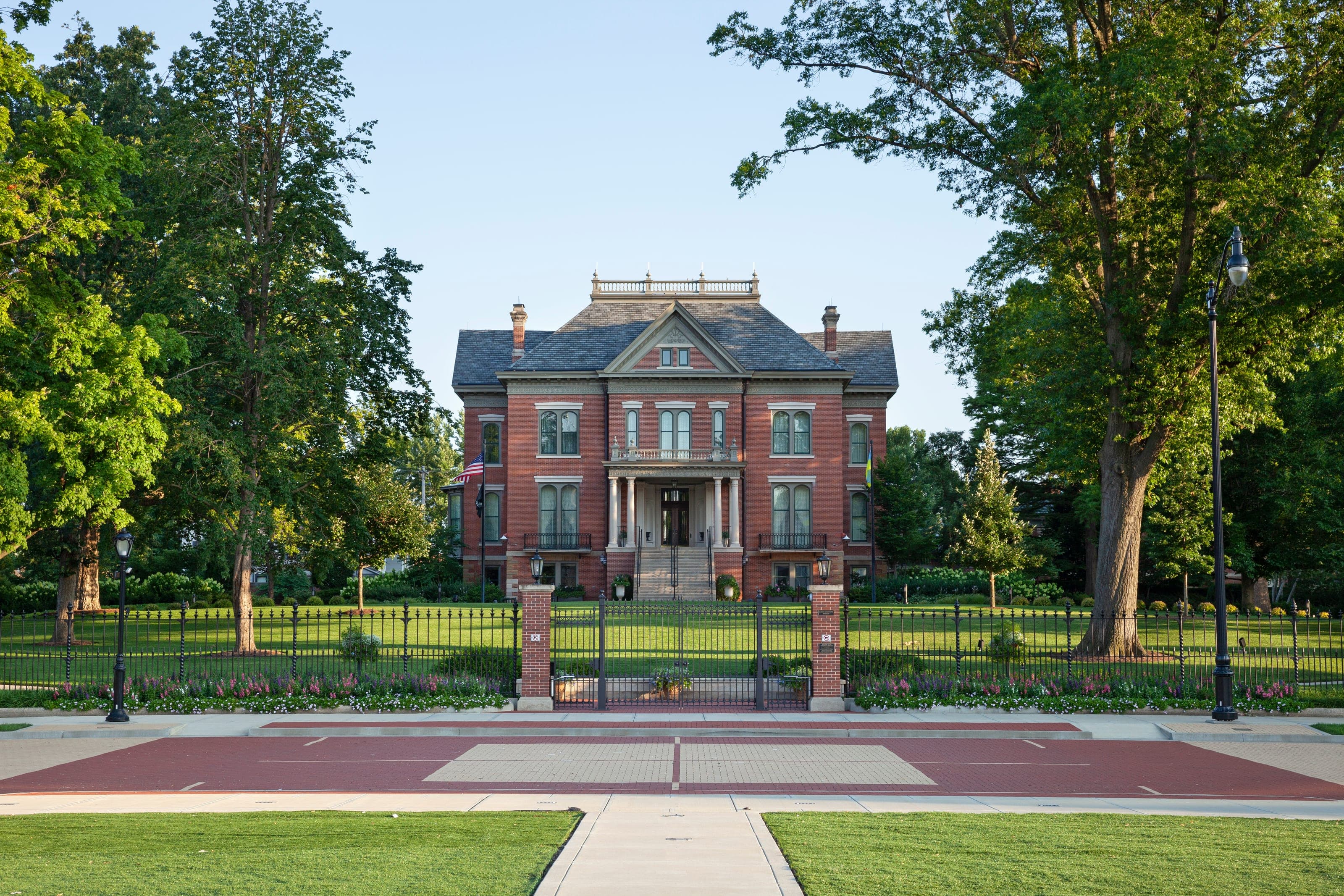 A red-brick mansion with a slate roof behind iron railings, framed by mature trees