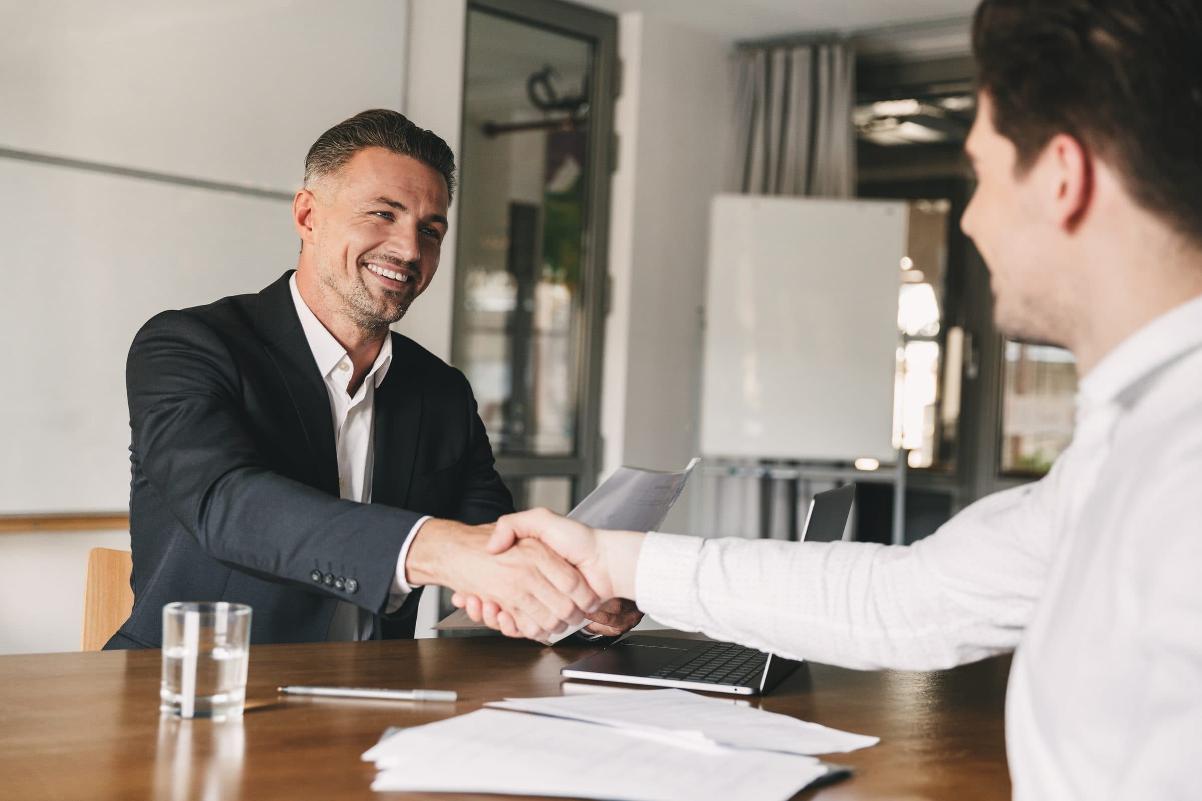 Two men in business dress smiling and shaking hands across a meeting table after an interview
