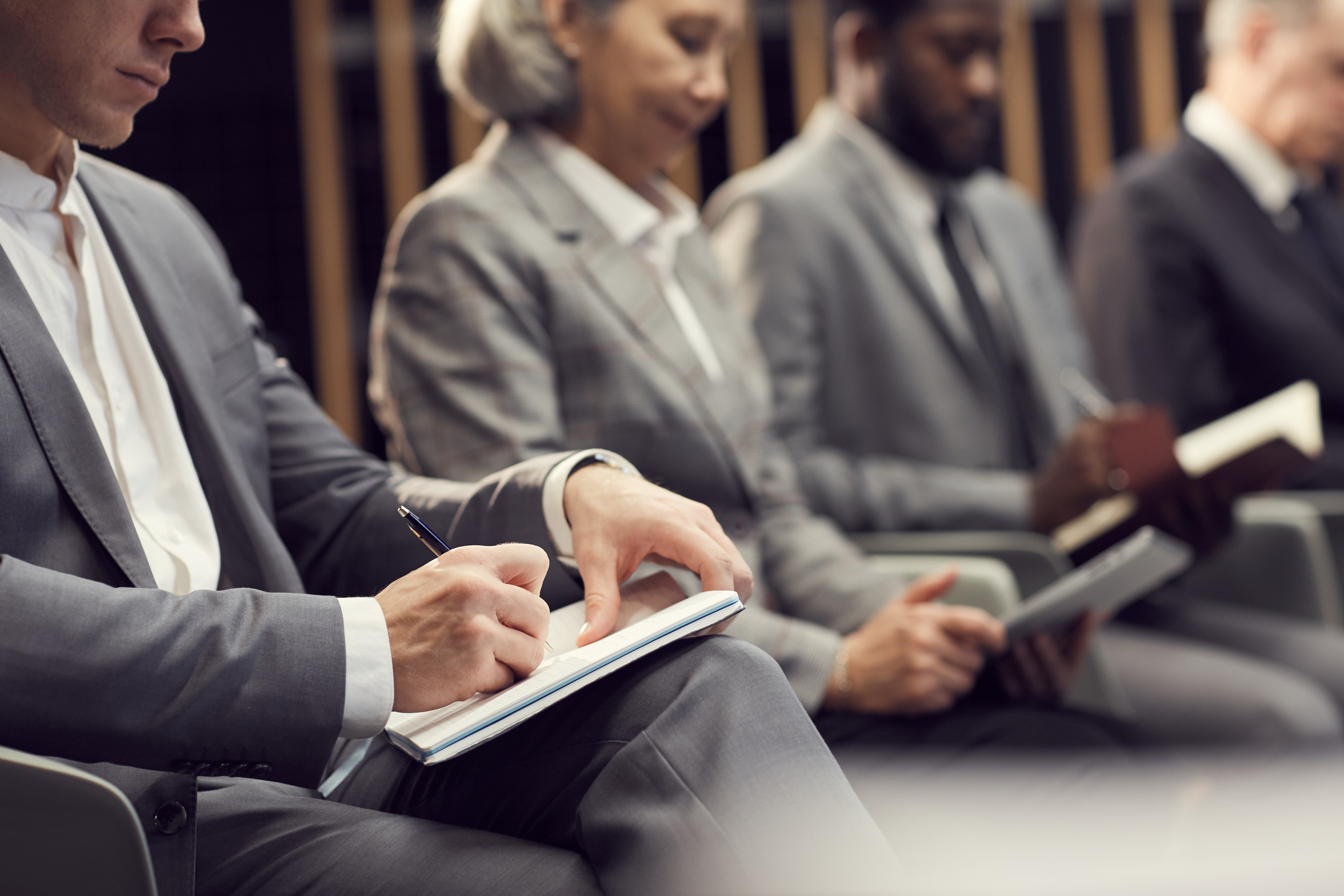 A person in a grey suit writing in a notebook, seated beside colleagues at a conference