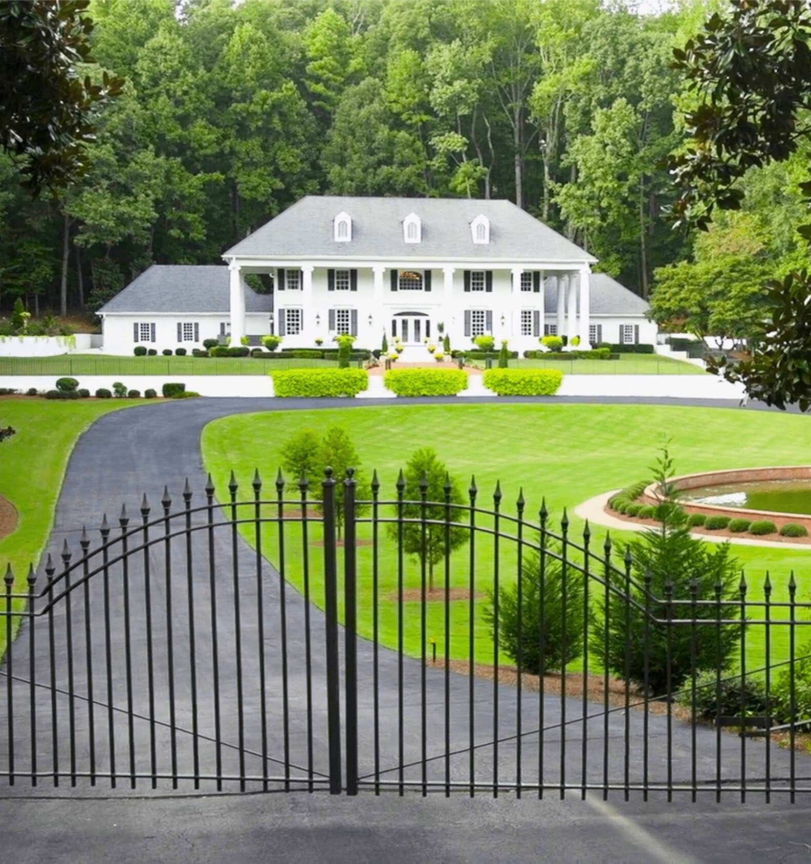 A white colonnaded country residence behind wrought-iron gates, clipped hedges and lawns in front of it and woodland rising behind